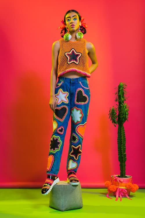 the model stands on a red background in a crochet orange tank top and blue jeans with black hearts and white stars from designer Katie Jones