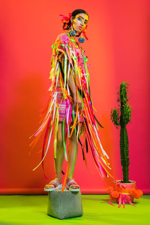 the model stands against a red background next to a cactus in a pink dress with many hanging colored ribbons from designer Katie Jones