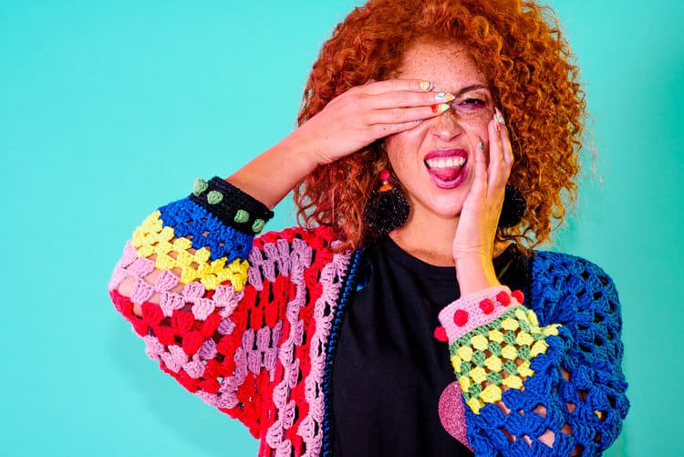 A girl on a green background in a colorful cardigan from designer Katie Jones grimaces at the camera
